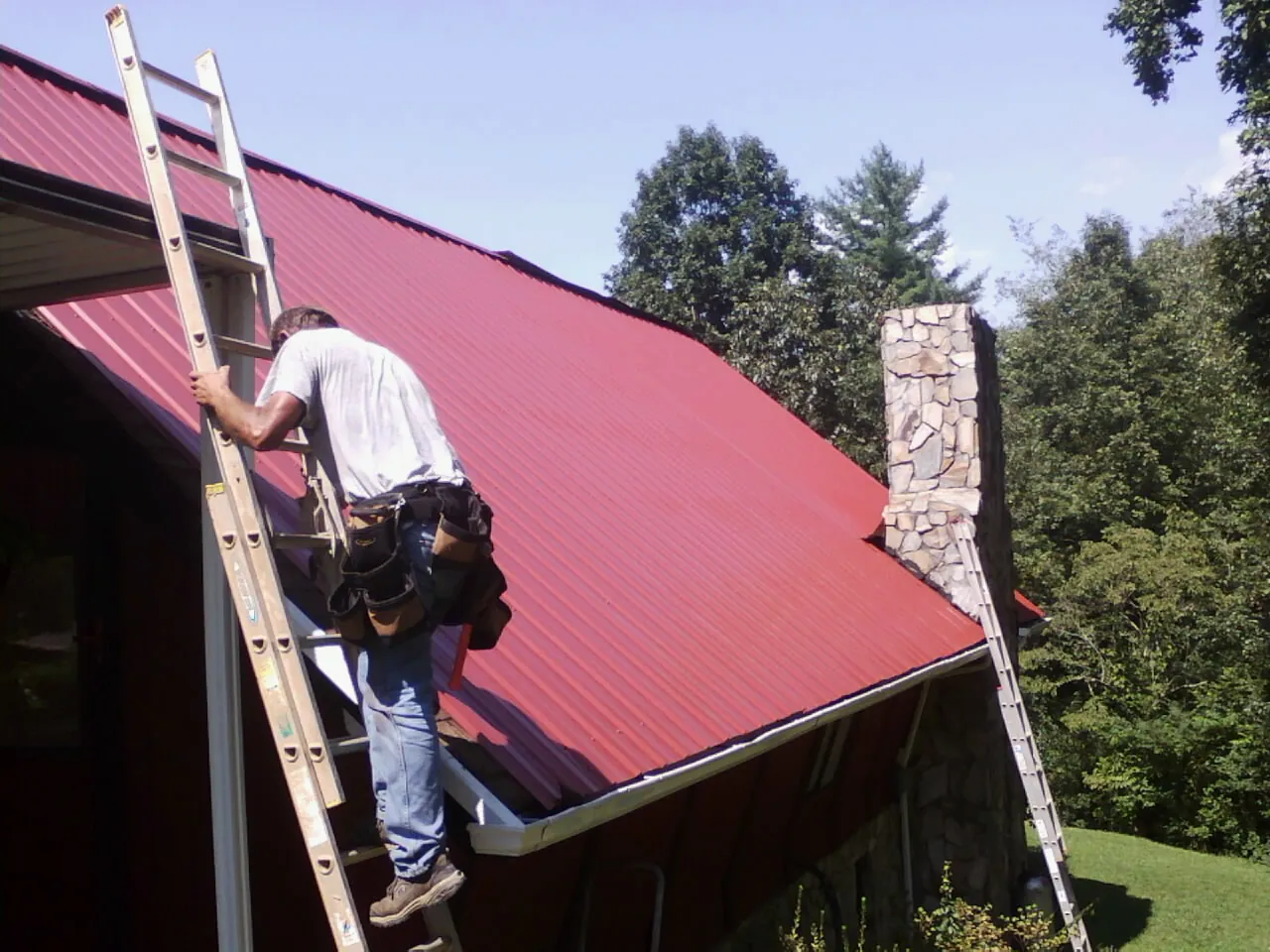 Roofer on a ladder beside a red metal roof and stone chimney — Soffit & Fascia Repair by Roofing Brothers in Landing